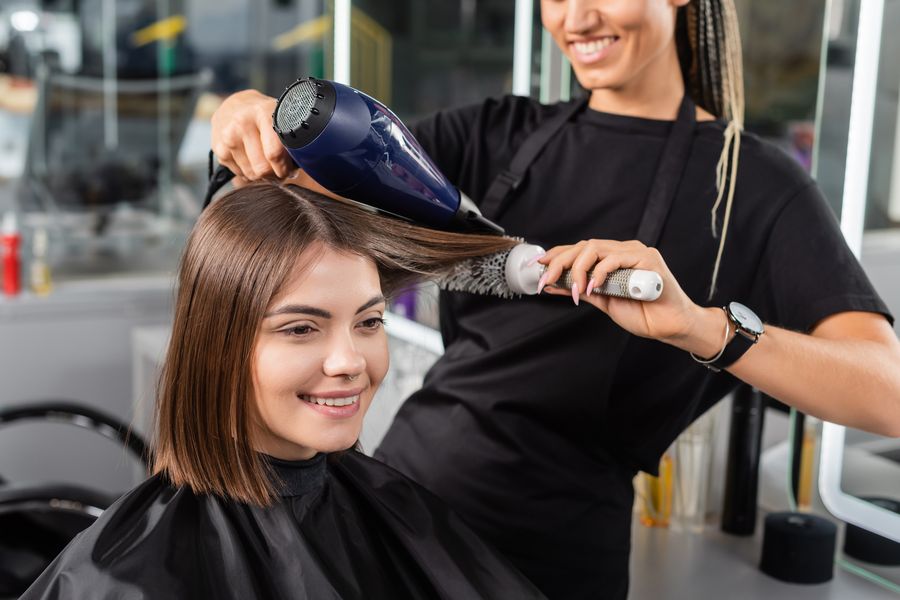 Stylist blow-drying a smiling client's shoulder-length brown hair with a round brush in a salon