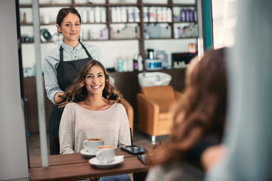 Happy client with fresh, bouncy waves and her smiling stylist reflected in the salon mirror, two cappuccinos on the counter