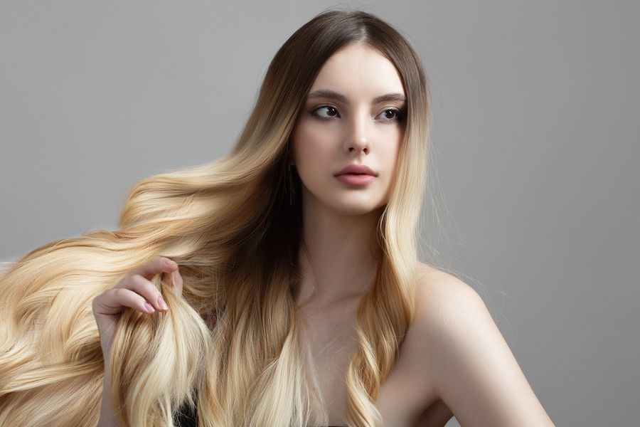 Young woman with long blonde balayage blending from darker roots to golden ends, gray background