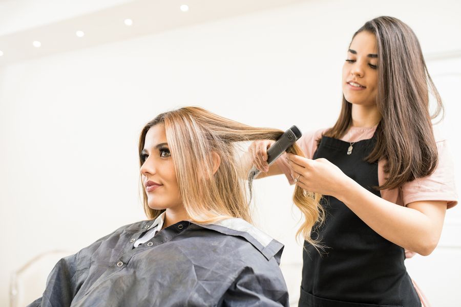 Stylist straightening a client's long highlighted hair with a flat iron in a bright salon